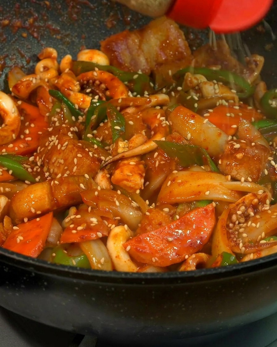 Eine Nahaufnahme von weißem Sesam, der aus einem Streuer mit roter Kappe auf eine glänzende Pfanne mit Tintenfisch, Schweinebauch, Karotten und grünen Chilis gestreut wird.
