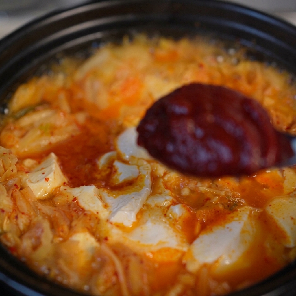 Ein Löffel mit einer Portion dicker, dunkelroter koreanischer Chilipaste über einer köchelnden Kimchi-Tofu-Suppe.