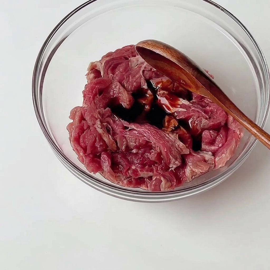 Dünn geschnittenes rohes Rindfleisch in einer Glasschüssel, das mit einem Holzlöffel mit dunklen Saucen vermengt wird.