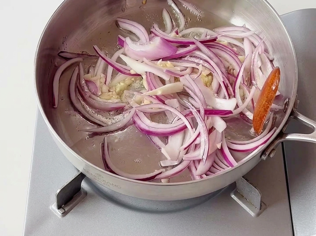 In Ringe geschnittene rote Zwiebeln und fein gehackter Knoblauch brodeln in einer flachen Edelstahlpfanne auf einem mobilen Kochfeld.