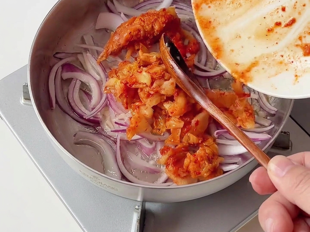 Eine Portion gehacktes rotes Kimchi wird von einem Teller in eine Edelstahlpfanne mit roten Zwiebelscheiben gegeben.