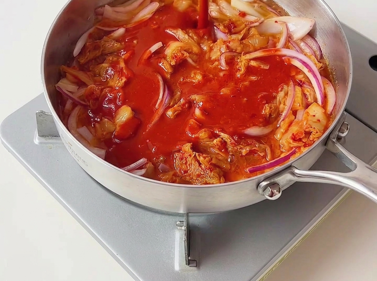Leuchtend rote Chilisauce wird über das angebratene Kimchi und die roten Zwiebeln in einer silbernen Pfanne gegossen.