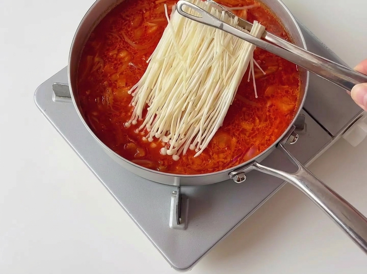 Eine Metallzange legt ein ordentliches Bündel weißer Enoki-Pilze in einen brodelnden roten Kimchi-Eintopf.