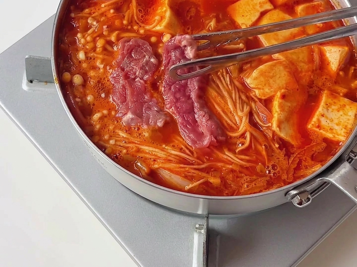 Eine Zange legt Stücke von rosafarbenem, mariniertem rohem Rindfleisch in einen heißen, brodelnden roten Eintopf neben Tofuscheiben.