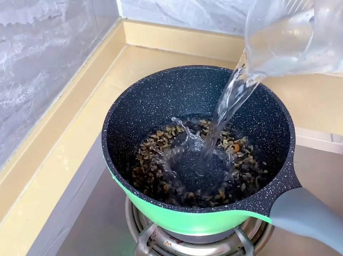 Klares Wasser wird aus einem Messbecher aus Kunststoff in einen grünen Topf auf einem Gasherd gegossen.