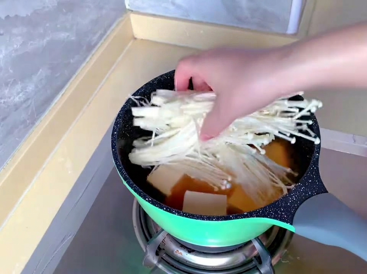 Eine Hand gibt ein Bündel langer, weißer Enoki-Pilze in einen Topf, in dem sich bereits Brühe und Tofu befinden.