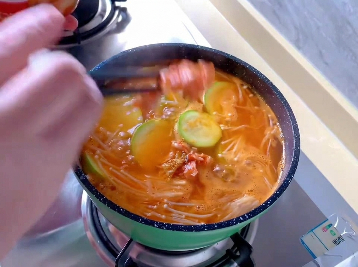 Rote Kimchi-Kohlstücke werden mit Essstäbchen in einen kleinen grünen Topf mit brodelnder orangefarbener Suppe, Enoki-Pilzen und Zucchinischeiben gegeben.