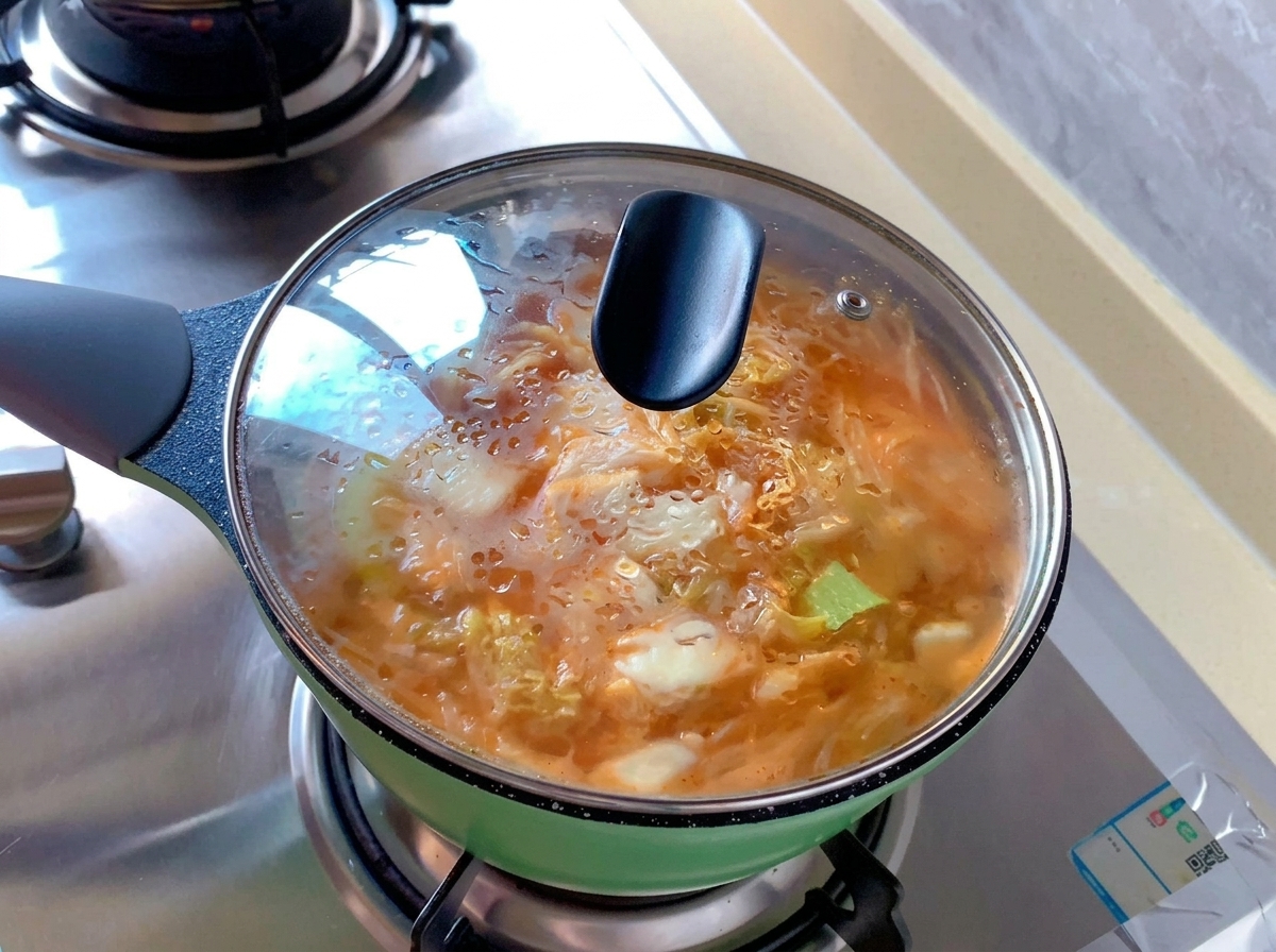 Ein kleiner grüner Topf mit orangefarbener Kimchi-Suppe, abgedeckt mit einem transparenten Glasdeckel, an dem Kondenswasser haftet.