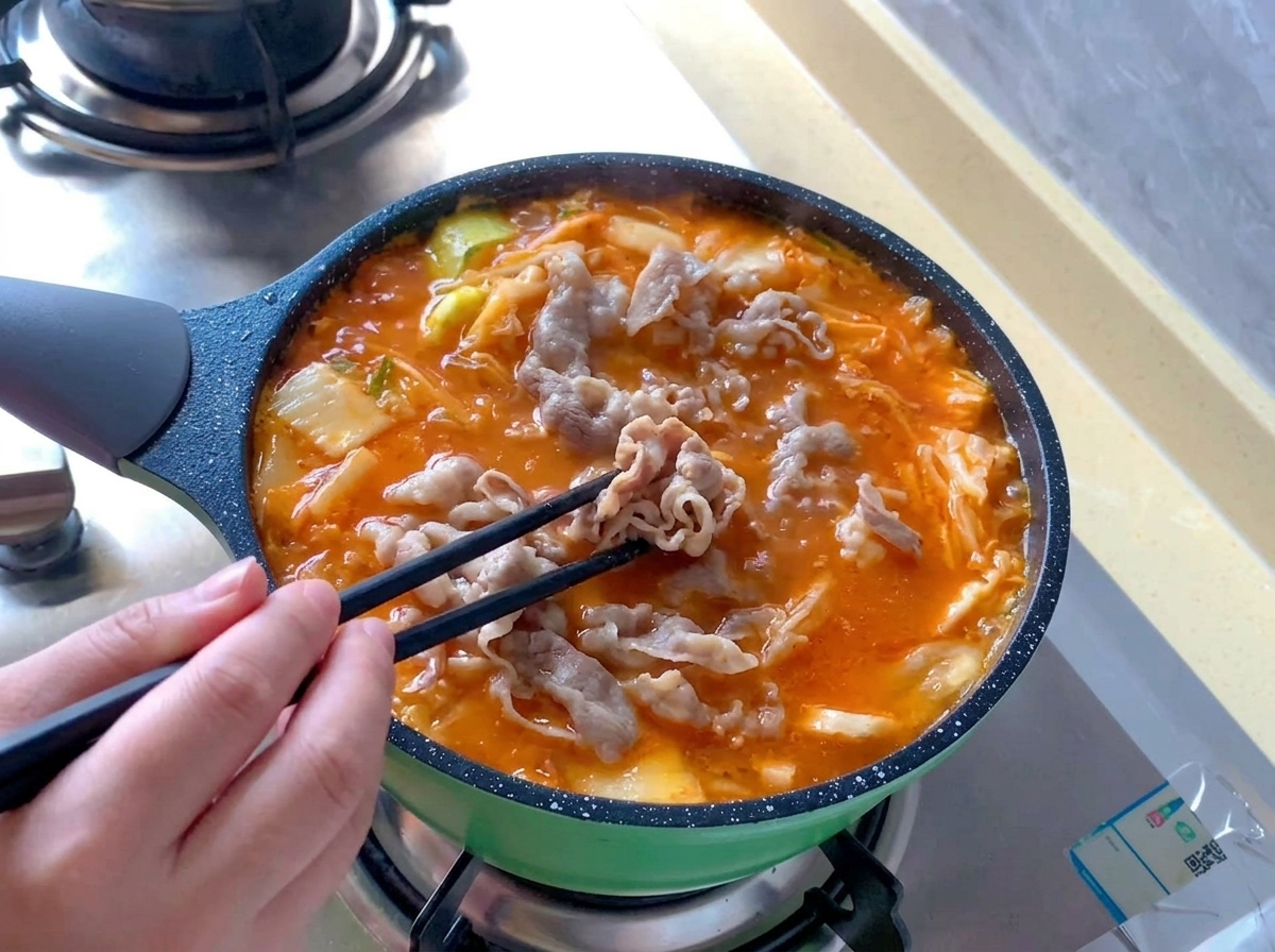 Blanchierte Rindfleischröllchen werden mit schwarzen Essstäbchen in einem kochenden Topf mit leuchtend rot-orangefarbener Kimchi-Suppe verteilt.