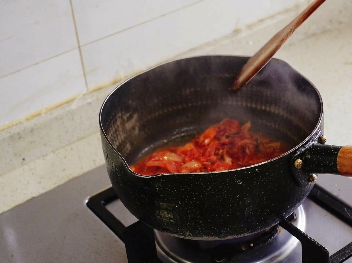 Kimchi wird mit einem Pfannenwender aus Holz in einem schwarzen Topf angebraten, Dampf steigt auf.