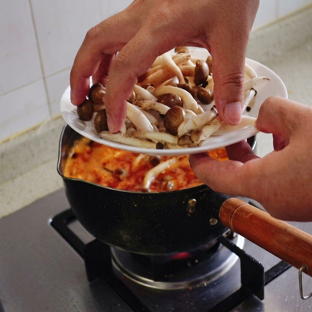 Hände geben eine Handvoll frischer Shimeji-Pilze mit braunen Kappen von einem weißen Teller in die kochende rote Suppe.