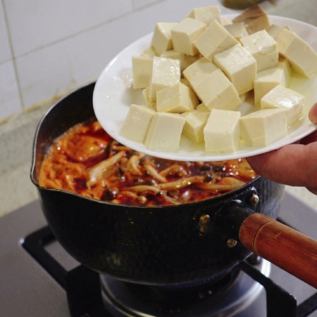 Eine Hand schiebt gleichmäßige Würfel aus weißem Seidentofu von einem weißen Teller in einen köchelnden Topf mit rotem Kimchi-Eintopf.