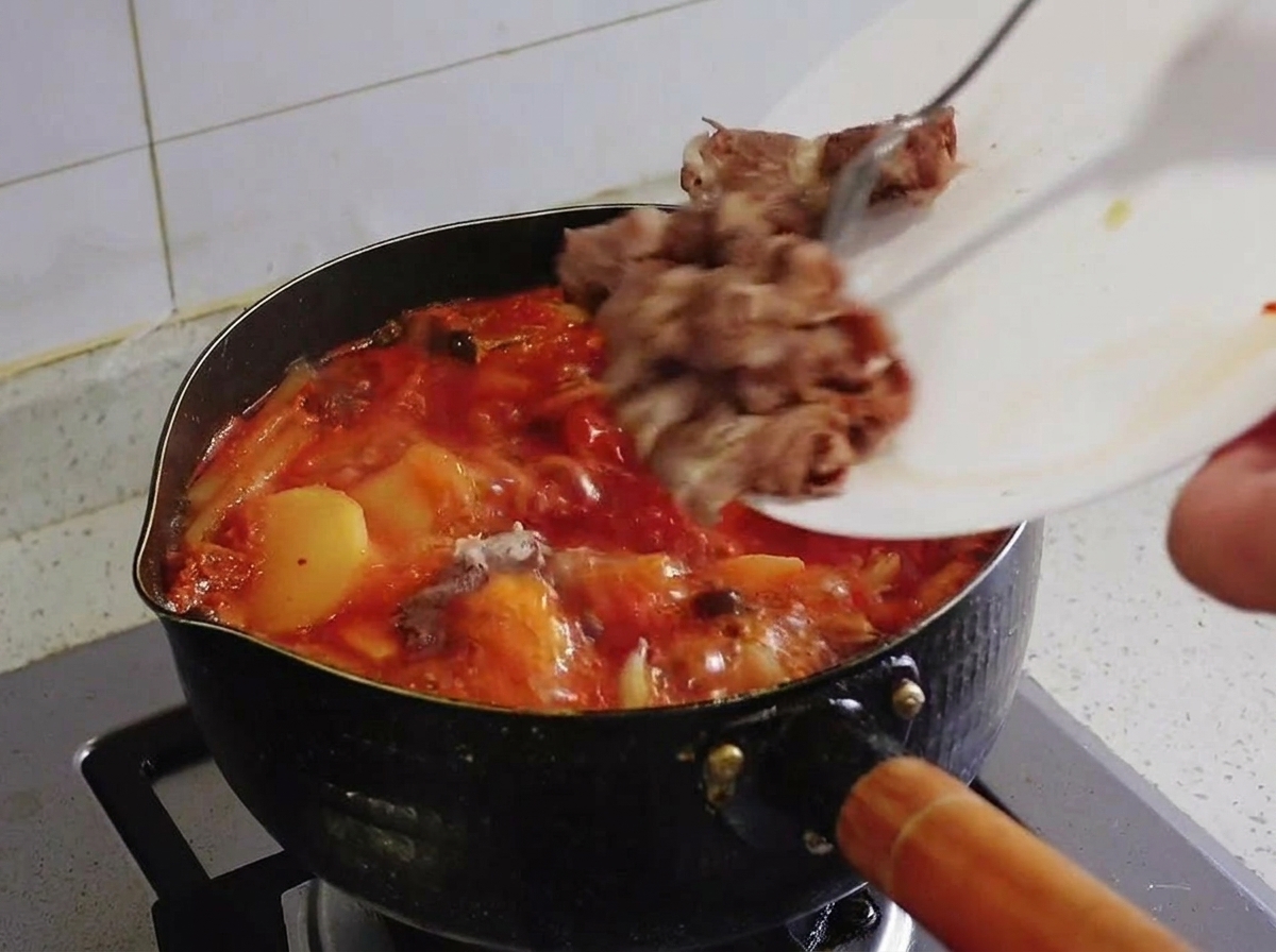 Blanchierte Rindfleischstücke werden von einem weißen Teller in einen kochenden Topf mit rotem Kimchi-Eintopf geschoben.