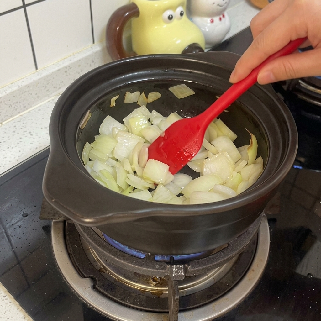 Ein roter Silikonspatel rührt geschnittene weiße Zwiebeln in einem schwarzen Tontopf über einer Gasflamme.