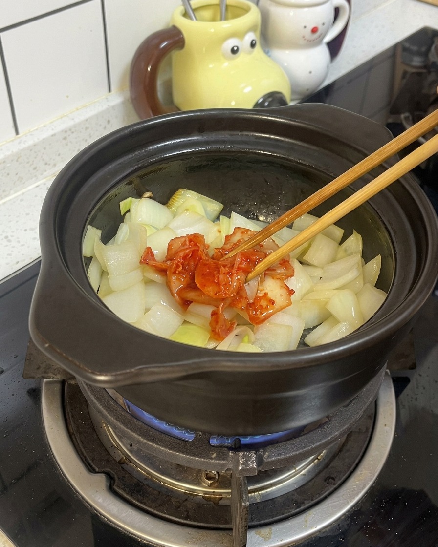 Holzstäbchen geben eine Portion rotes, fermentiertes Kimchi zu den angeschwitzten Zwiebeln in den schwarzen Tontopf.