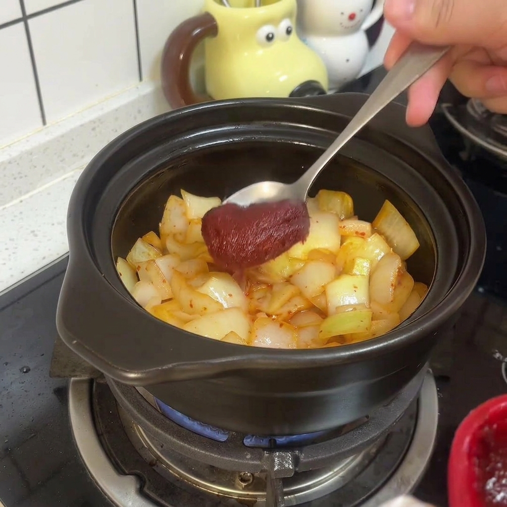 Ein Metalllöffel rührt dicke rote koreanische Chilipaste unter das angebratene Kimchi und die Zwiebeln im Tontopf.