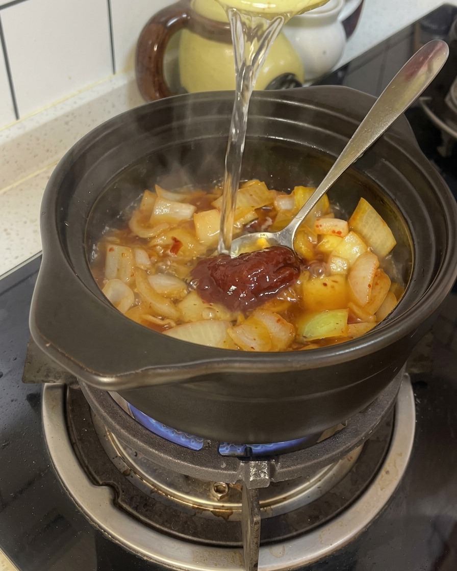 Wasser wird in einen schwarzen Tontopf gegossen, der bereits die gewürzten Zwiebeln und die rote Chilipaste enthält.
