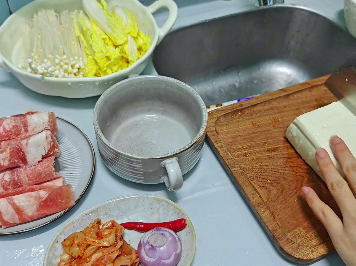 Nahaufnahme einer Hand, die mit einem Küchenmesser einen großen Block weißen Tofu in dicke, rechteckige Stücke auf einem Holzschneidebrett schneidet.