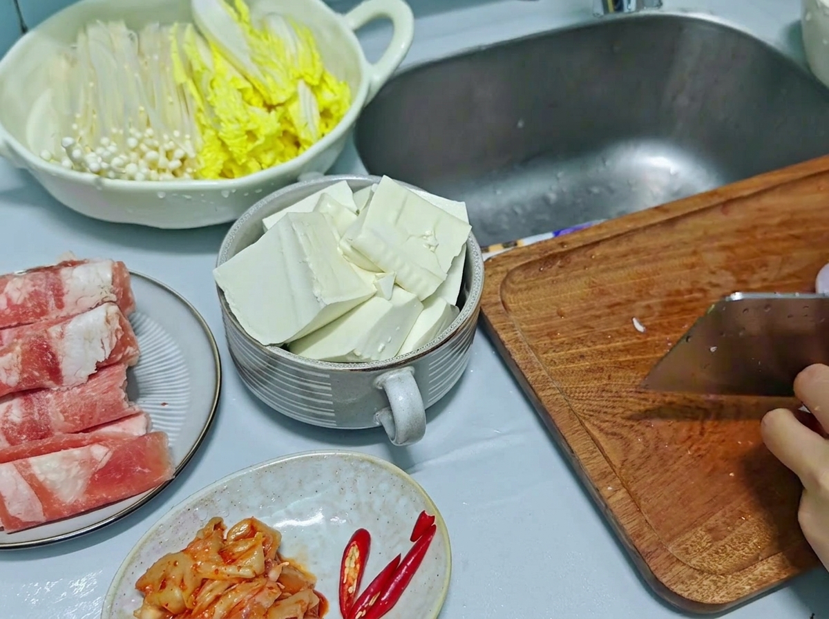 Ein Hackmesser hackt rote Zwiebeln und rote Chilischoten auf einem Holzschneidebrett, im Hintergrund steht geschnittener Tofu in einer Schale.