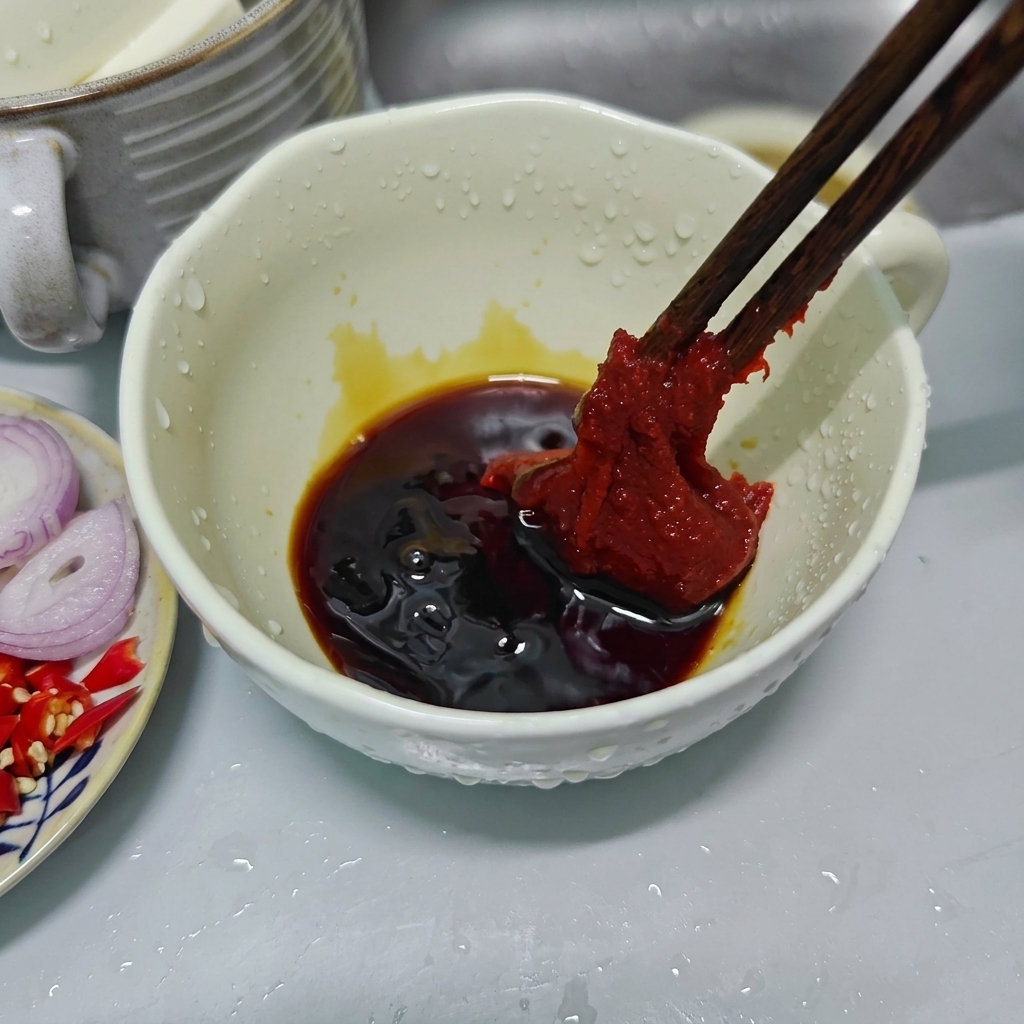 Holzstäbchen halten einen großen Klecks dicke, rote koreanische Chilipaste über einer weißen Keramikschale mit dunklen Saucen.