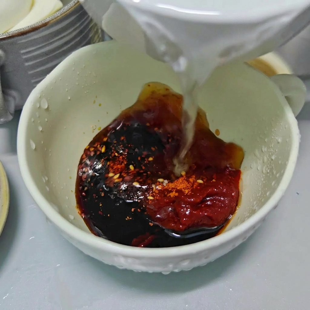 Klares Wasser wird in eine weiße Schale gegossen, die mit dunkler Sojasauce, roter Chilipaste und Chiliflocken gefüllt ist.