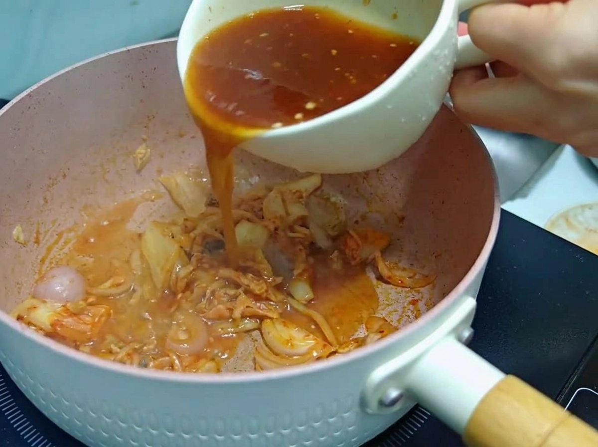 Eine Person gießt eine rötlich-braune Saucenmischung aus einer weißen Tasse in eine rosa Pfanne mit angebratenem Kimchi und Zwiebeln.