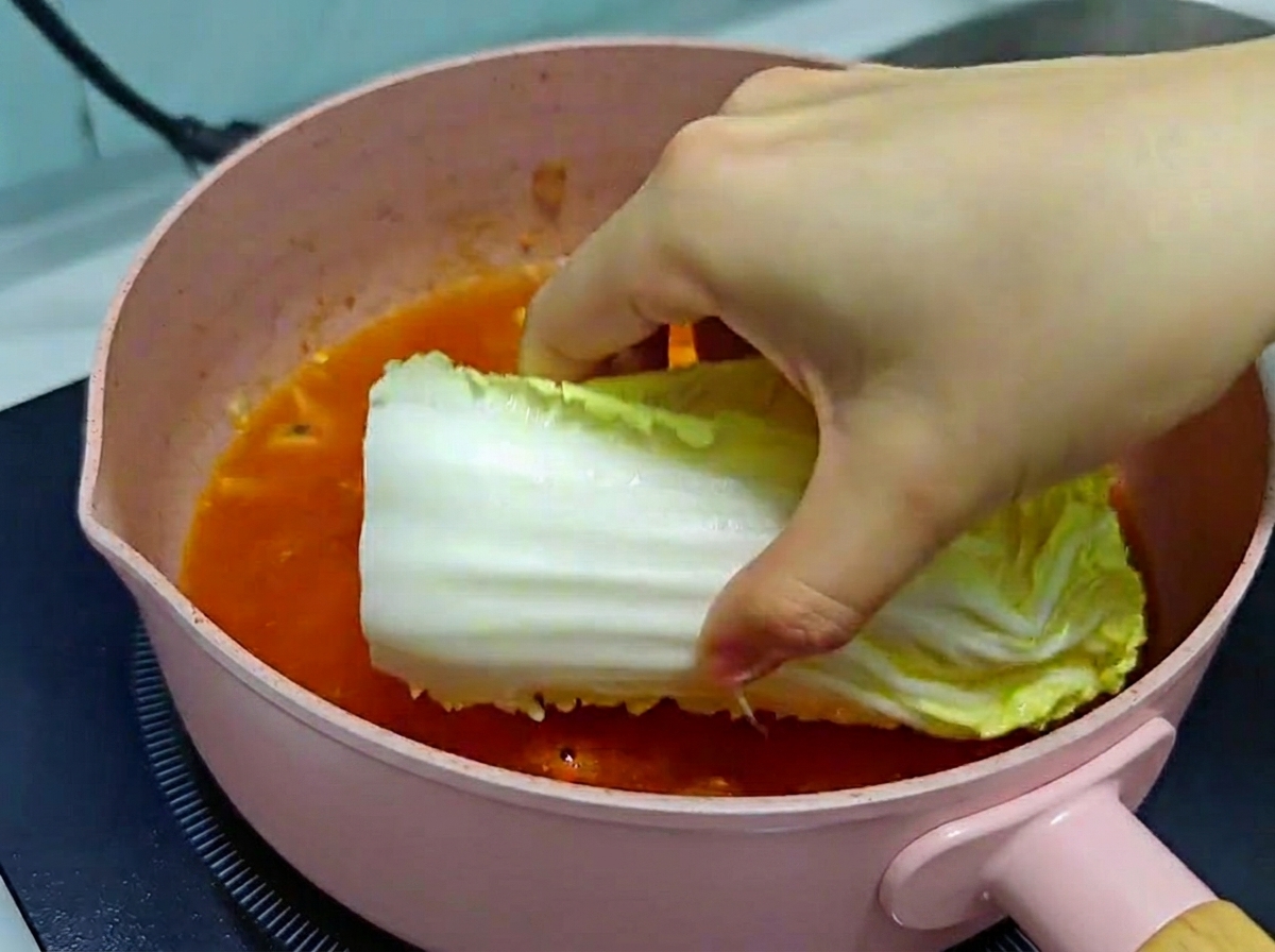 Eine Hand legt vorsichtig ein großes Stück frischen Chinakohl in eine rosa Pfanne mit köchelnder roter Kimchi-Suppenbrühe.