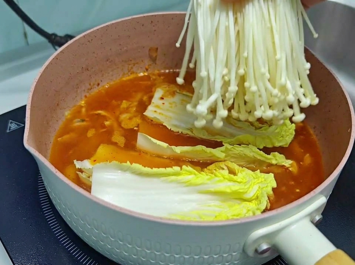 Nahaufnahme einer Hand, die ein frisches, sauberes Bündel weißer Enoki-Pilze in einen rosa Kochtopf mit einer reichhaltigen, orange-roten Kimchi-Suppenbasis und Chinakohl legt.
