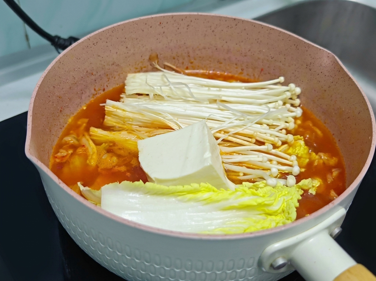 Ein großer Block frischer weißer Tofu liegt auf den Enoki-Pilzen und dem Kohl in einem Topf mit orangefarbener Kimchi-Suppe.