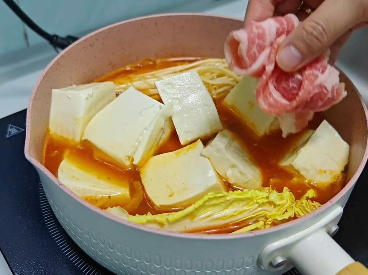 Eine Hand schichtet dünne Röllchen aus rohem Rindfleisch vorsichtig über Tofuwürfel und Gemüse in einem köchelnden Kimchi-Suppentopf.