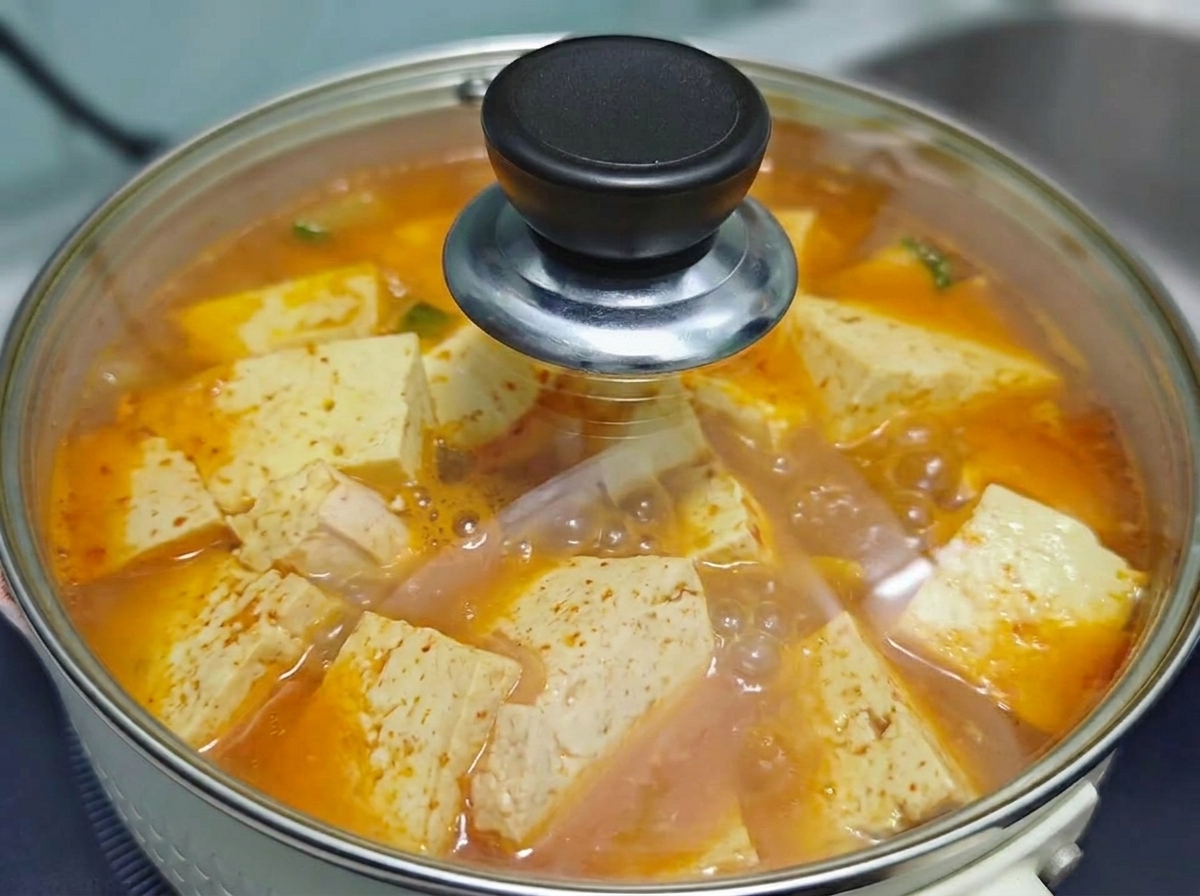 Ein klarer Glasdeckel bedeckt den Topf mit der kochenden koreanischen Kimchi-Rindfleisch-Tofu-Suppe, während sie auf dem Herd köchelt.