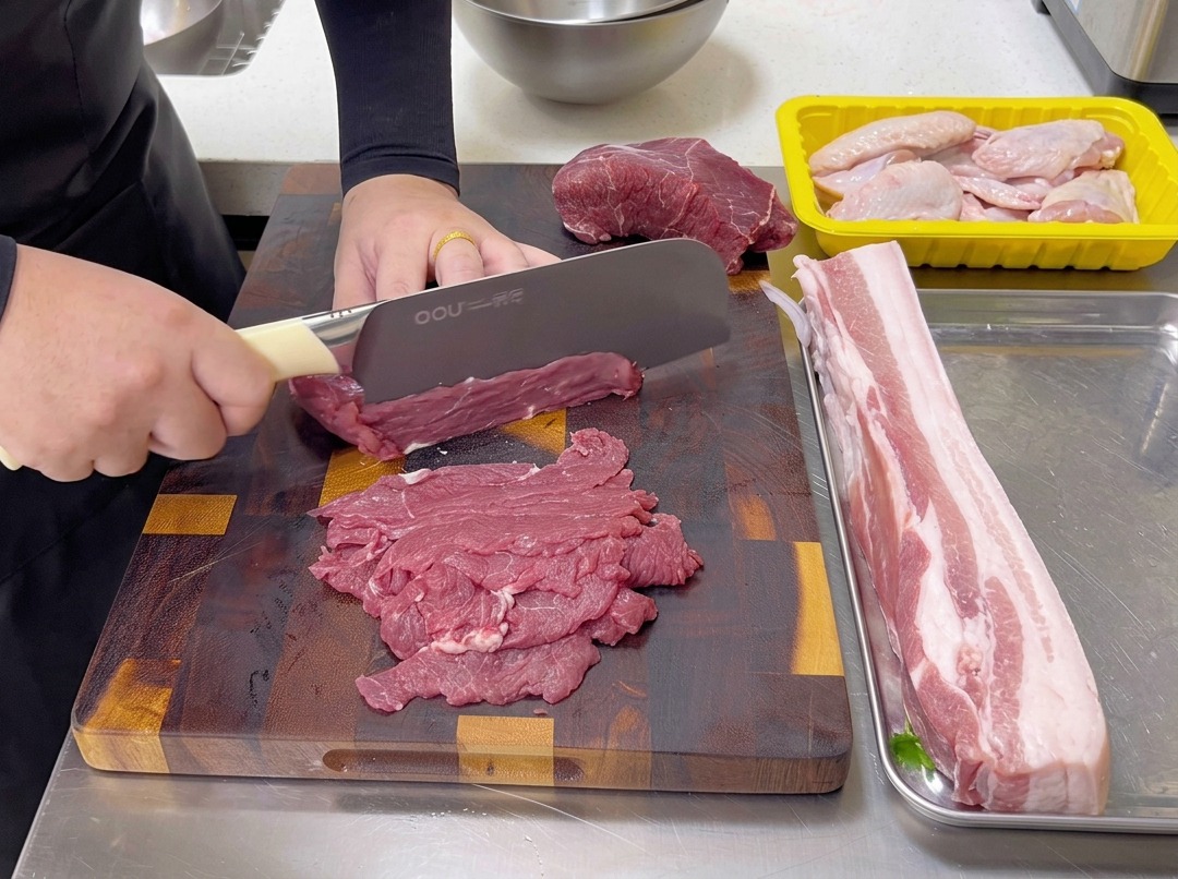 Rohes Rindfleisch wird mit einem Kochmesser auf einem Holzbrett quer zur Faser in dünne Scheiben geschnitten.