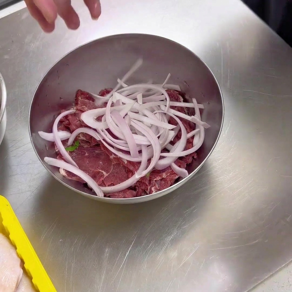 Dünne Scheiben von rohem Rindfleisch in einer Metallschüssel, garniert mit einer Handvoll geschnittener weißer Zwiebeln.
