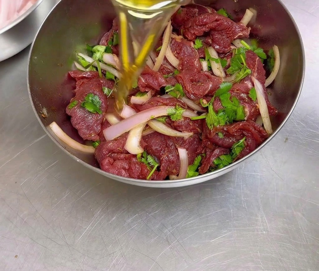 Speiseöl wird in eine Edelstahlschüssel mit mariniertem Rindfleisch, Zwiebelscheiben und Koriander gegossen.