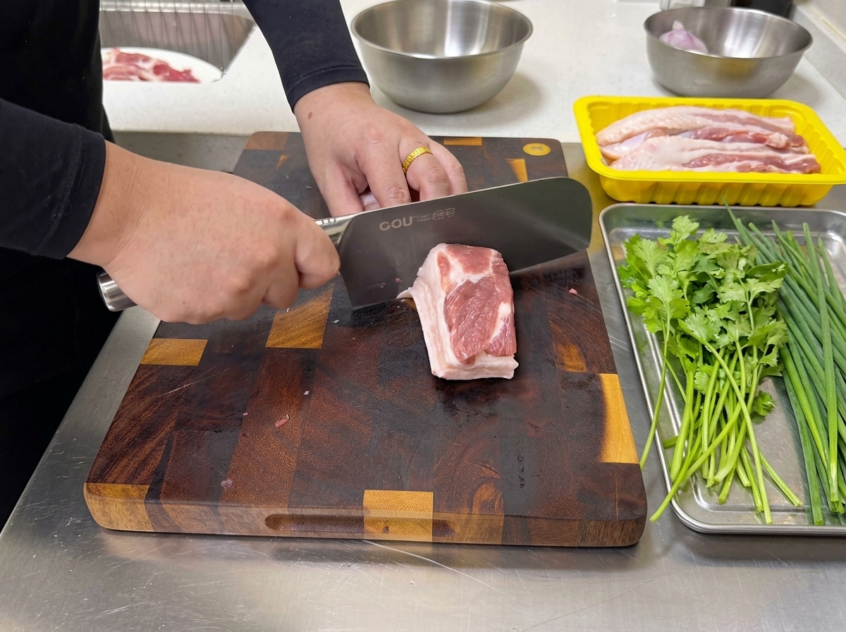 Ein großes Küchenmesser schneidet ein Stück rohen, geschichteten Schweinebauch in dünne Streifen auf einem Schneidebrett.