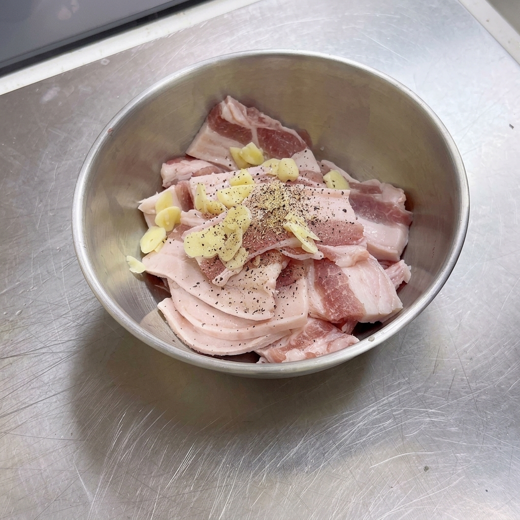 Eine Edelstahlschüssel gefüllt mit rohen Schweinebauchscheiben, Knoblauchstücken und frisch gemahlenem schwarzen Pfeffer.