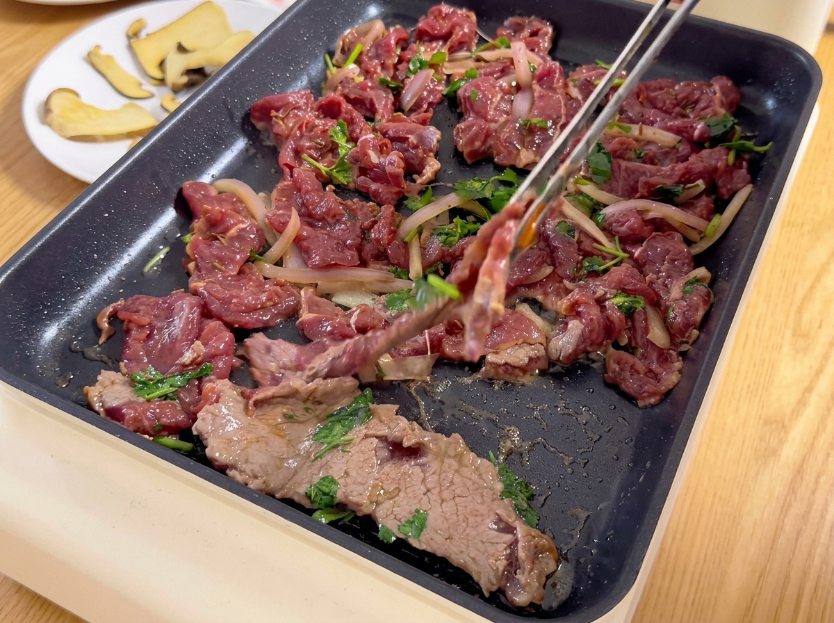 Dünn geschnittenes mariniertes Rindfleisch mit Zwiebeln und Koriander wird mit einer Zange auf einen heißen Elektrogrill gelegt.