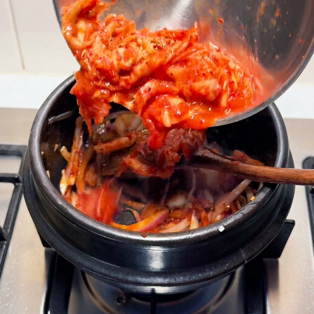 Leuchtend rotes, gehacktes saures Kimchi wird aus einer Edelstahlschüssel in einen schwarzen Steintopf auf dem Herd gegossen.
