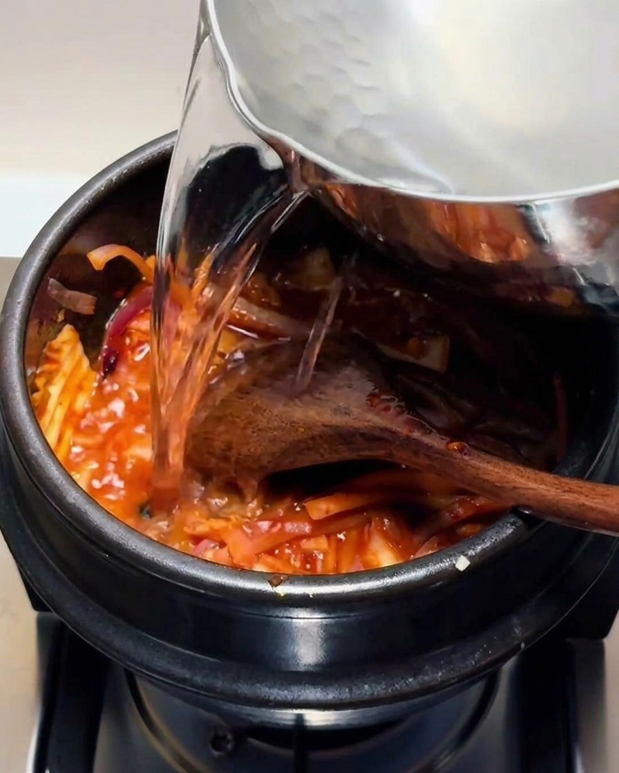 Klares heißes Wasser wird aus einem Metalltopf in einen schwarzen Steintopf mit der roten Kimchi-Mischung gegossen.