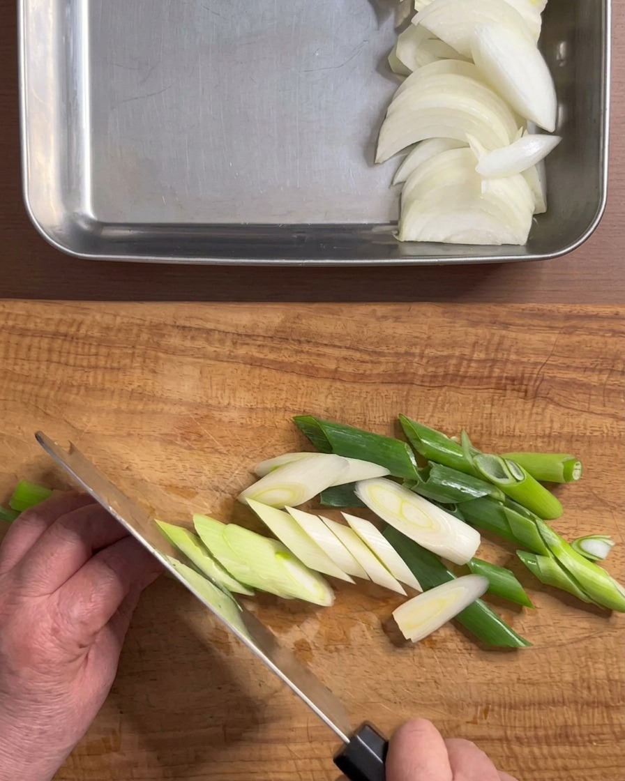 Hände schneiden eine frische Frühlingszwiebel schräg auf einem Holzschneidebrett.