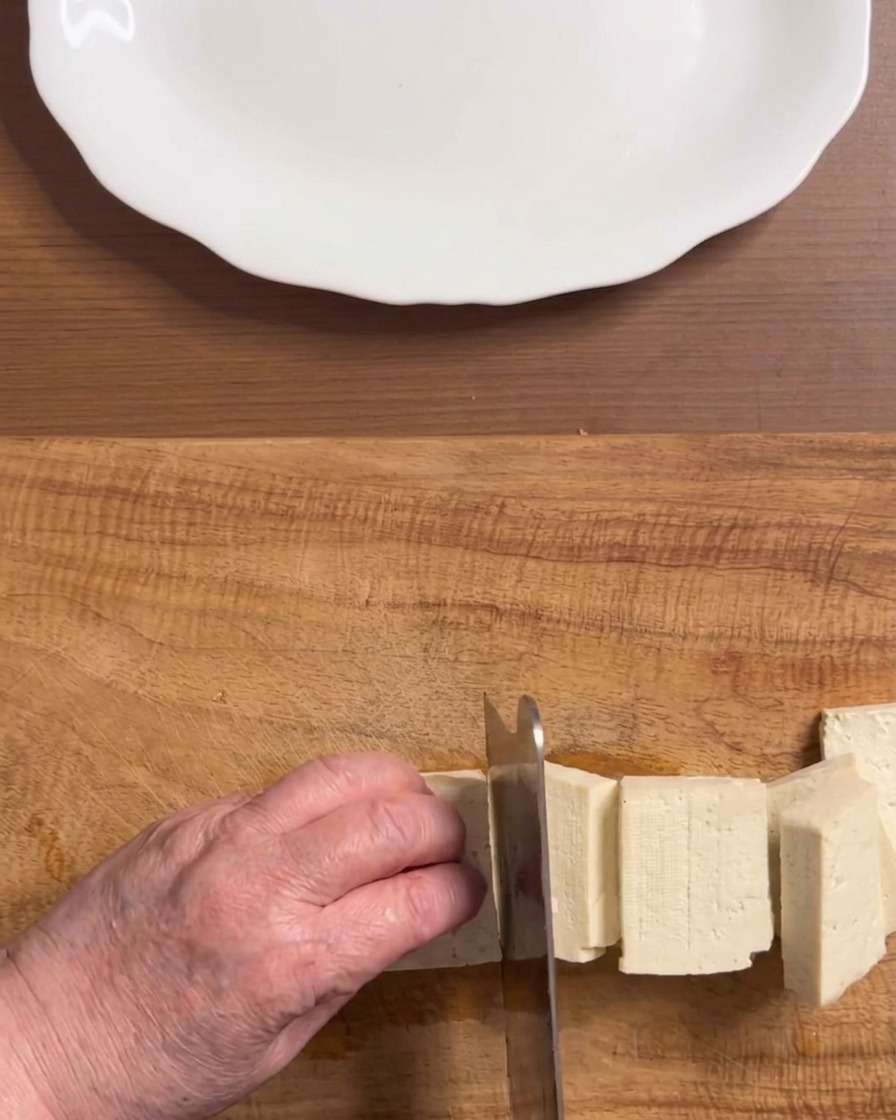 Hände schneiden einen Block festen weißen Tofu auf einem Holzschneidebrett in gleichmäßige rechteckige Stücke.