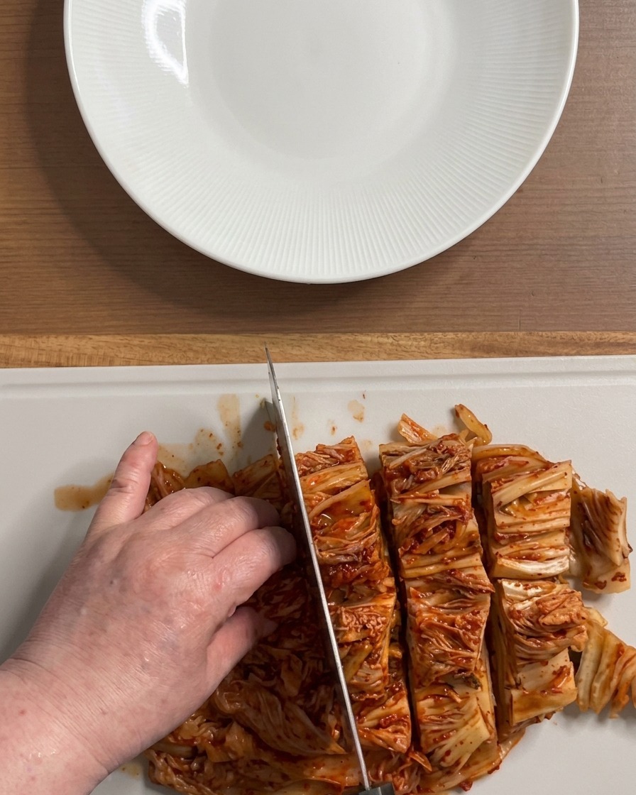 Hände schneiden einen großen, leuchtend roten Block fermentiertes Kimchi auf einem weißen Schneidebrett in mundgerechte Stücke.