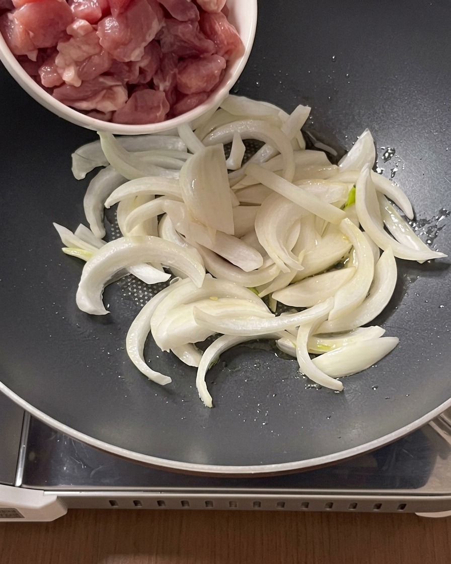 Rohe Schweinefleischstücke werden aus einer kleinen Schüssel in eine Pfanne mit geschnittenen Zwiebeln und Öl gegeben.