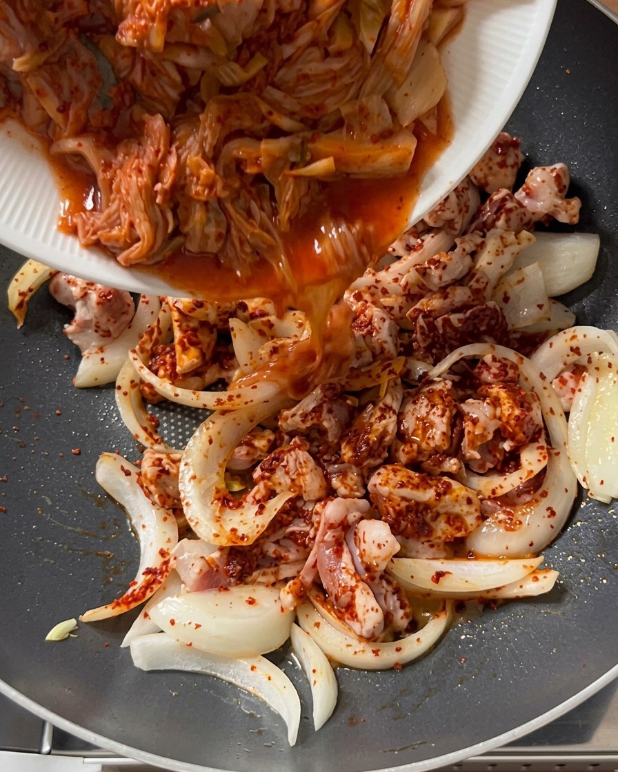 Gehacktes Kimchi wird von einem weißen Teller in eine Pfanne mit gewürztem Schweinefleisch und Zwiebeln gegossen.
