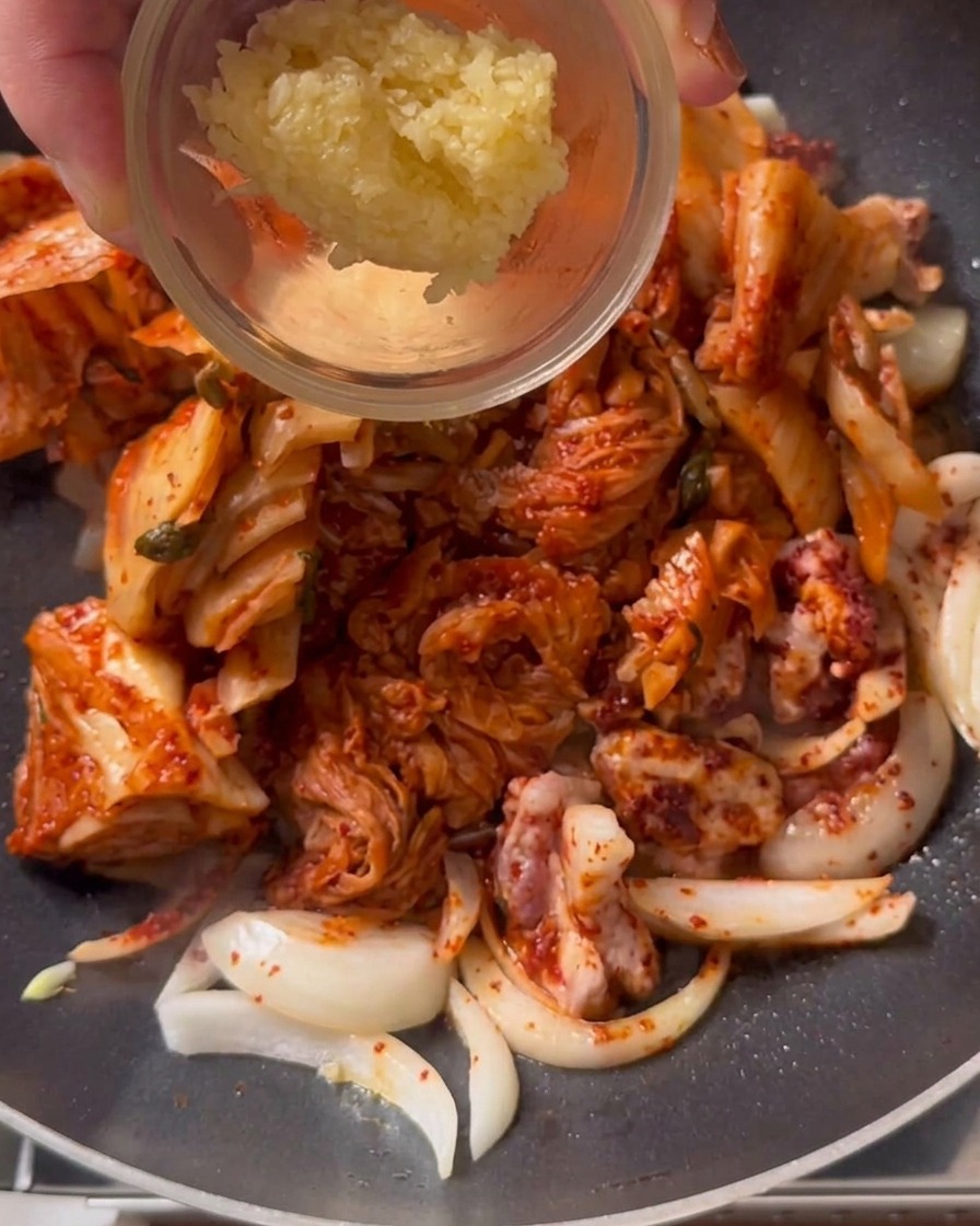 Ein Löffel gehackter Knoblauch wird in eine Pfanne mit anbratendem Kimchi und Schweinefleisch gegeben.