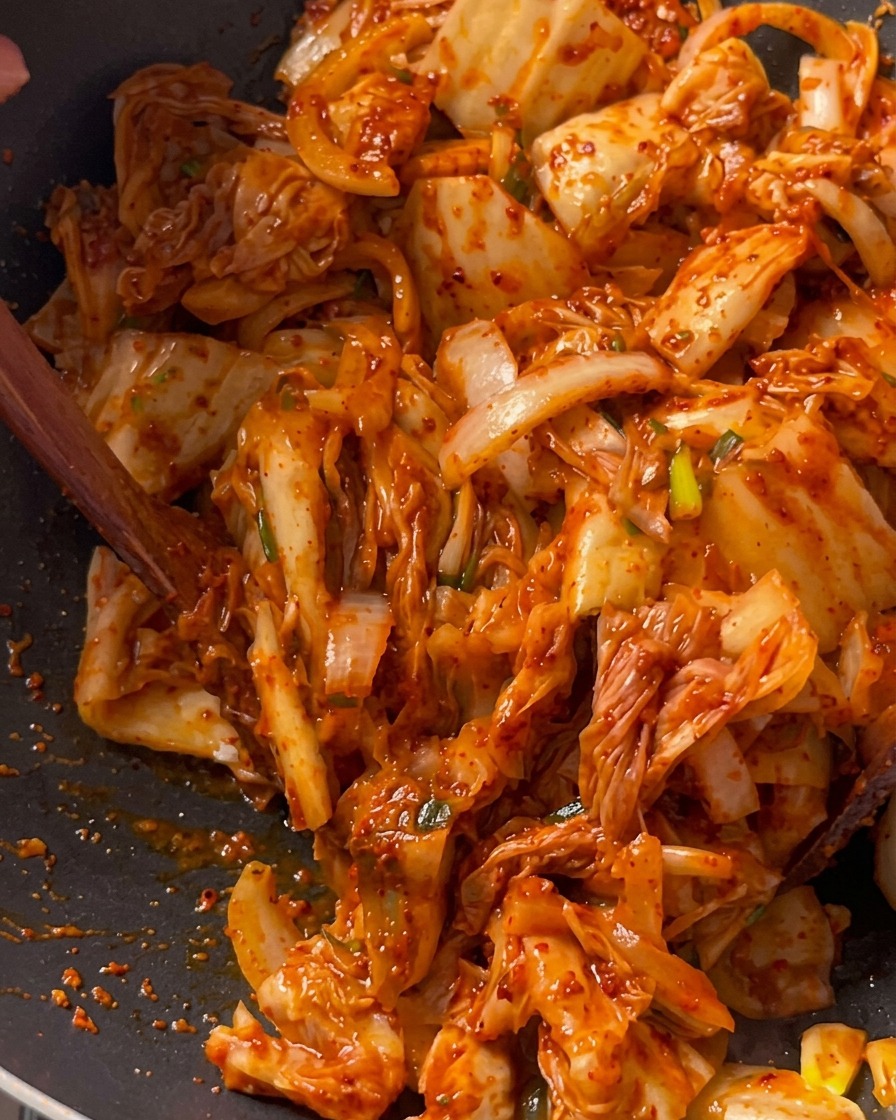 Kimchistücke werden zusammen mit Schweinefleisch und Zwiebeln in einem tiefen schwarzen Wok mit einem Holzspatel angebraten.