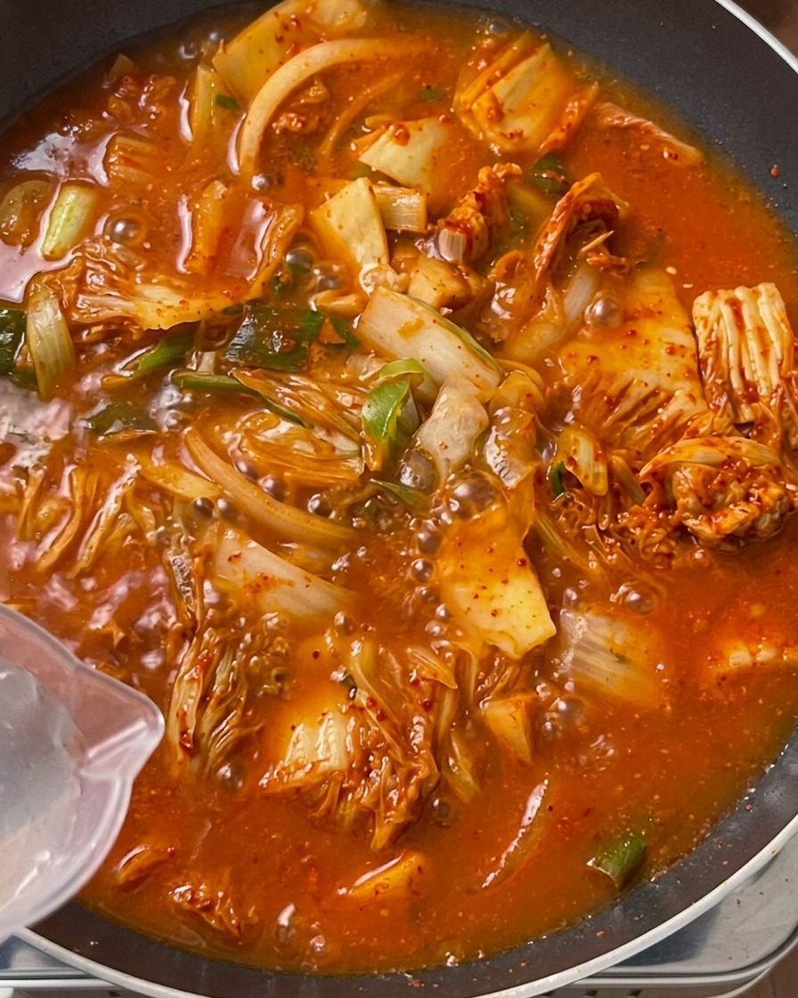 Klares Wasser wird aus einem transparenten Plastikkrug in eine große Pfanne gegossen, die mit einer sprudelnden, leuchtend orange-roten Kimchi-Fleisch-Mischung gefüllt ist.