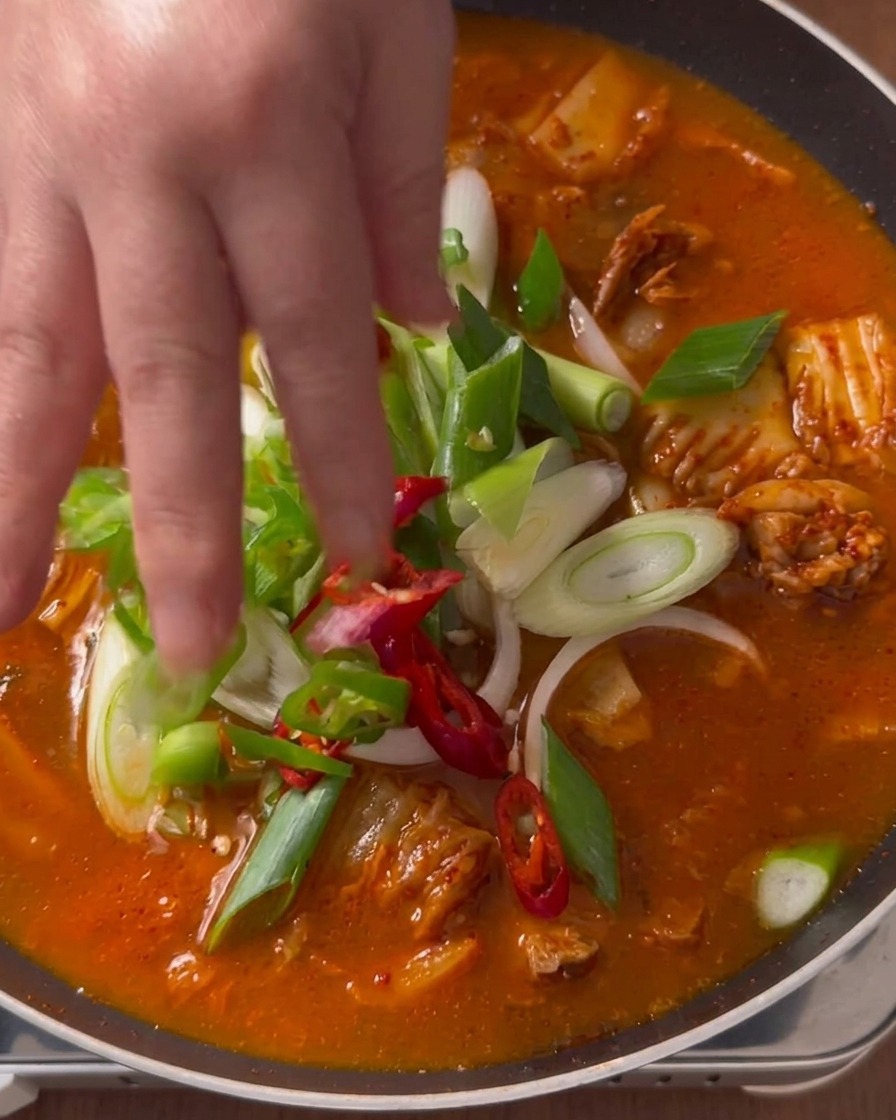 Eine Hand legt frisch geschnittene Frühlingszwiebeln, grüne Chilis und rote Paprikascheiben auf einen köchelnden roten Kimchi-Eintopf in einer Pfanne.