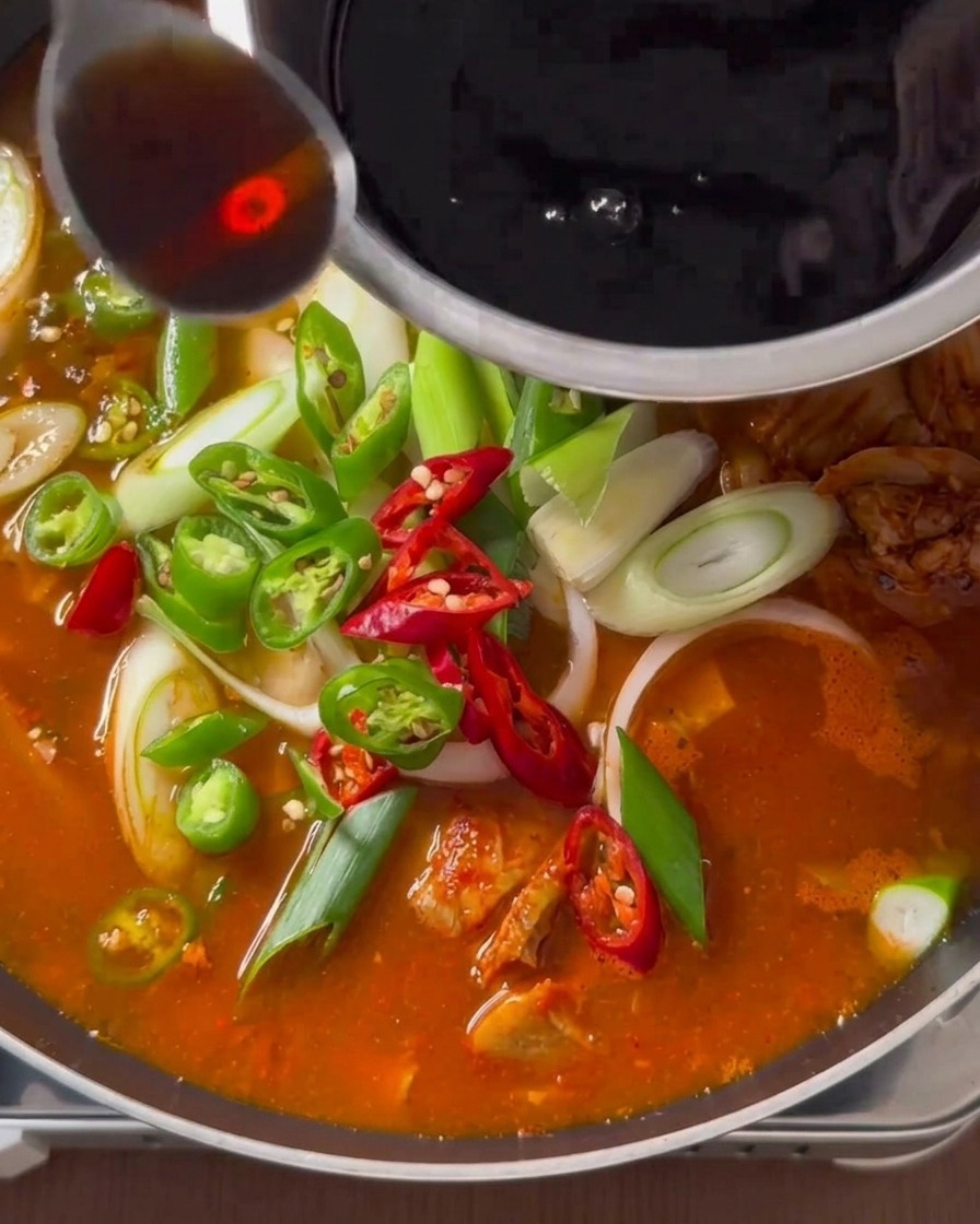 Eine dunkle flüssige Würze wird aus einer Edelstahlschüssel über einen köchelnden Kimchi-Eintopf gelöffelt, der mit frischen Paprika und Frühlingszwiebeln belegt ist.