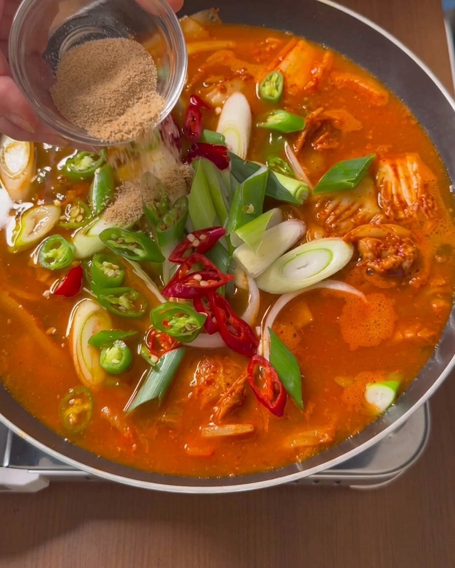 Trockenes braunes Rinderwürzpulver wird aus einer kleinen klaren Glasschale auf die Oberfläche eines köchelnden roten Eintopfs gestreut, der mit Paprika und Frühlingszwiebeln gefüllt ist.